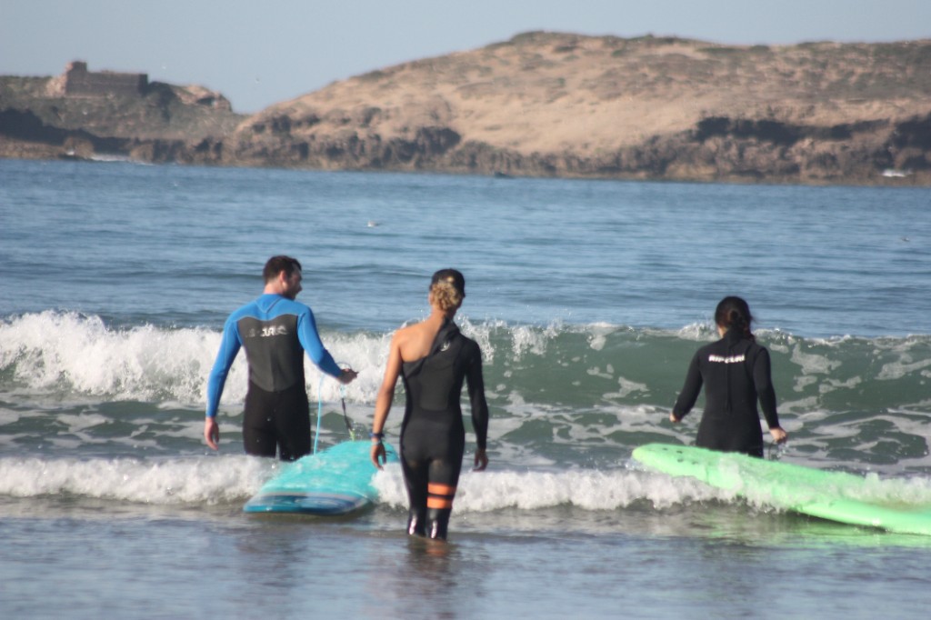 Tres surfistas con neopreno entrando al Atlántico con tablas soft-top; isla rocosa al fondo — Essaouira, Marruecos