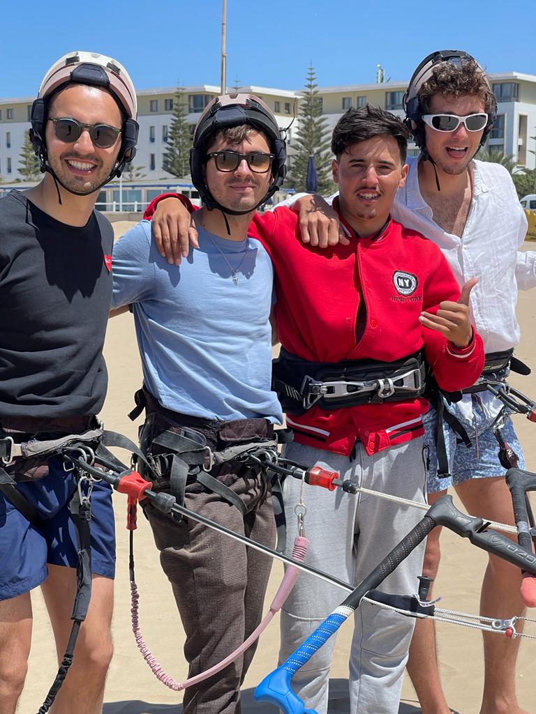 Cuatro hombres en una playa soleada de Essaouira, arneses de kite, barras y líneas en primer plano — cascos, gafas de sol, poses relajadas y shaka, edificio blanco y cielo azul al fondo.