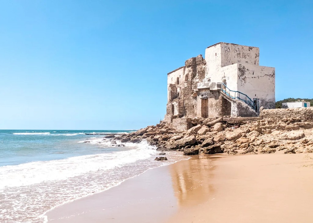 Vista de la costa de Sidi Kaouki con playa y edificio antiguo sobre rocas.