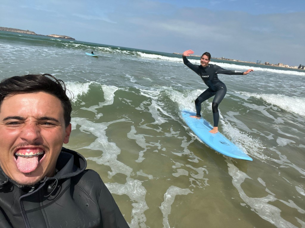 Selfie en el agua durante una clase de surf en Essaouira — alumna sonriendo de pie sobre tabla azul en la espuma, monitor con neopreno con capucha en primer plano, costa al fondo.