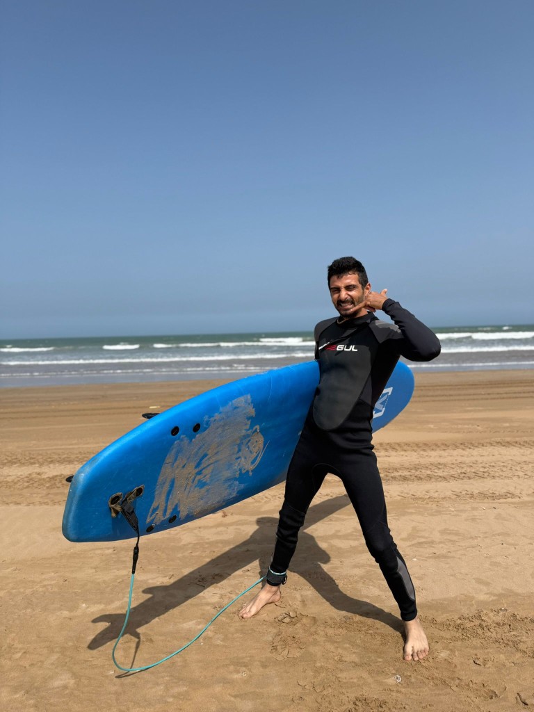 Monitor sonriente en neopreno en la playa, tabla azul bajo el brazo, shaka — escuela surf Essaouira.