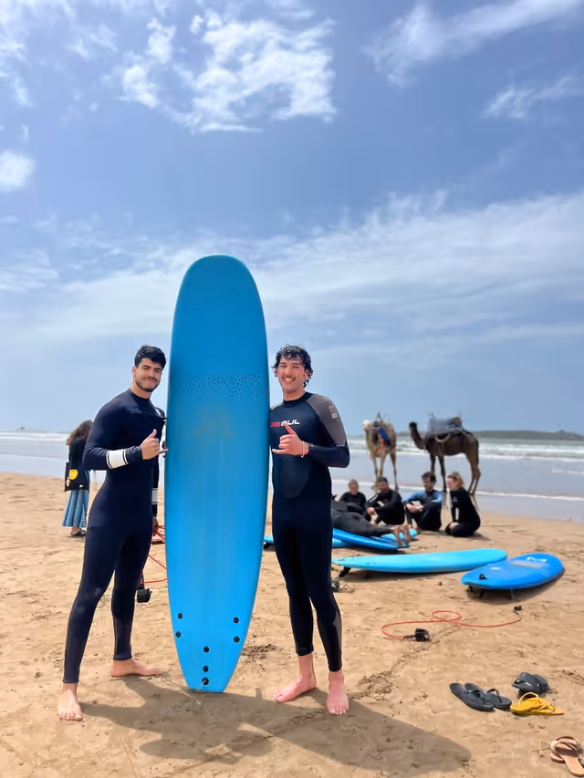 Clases de surf en la playa de Essaouira: dos monitores en neopreno, tabla azul, shaka, grupo y camellos cerca del mar