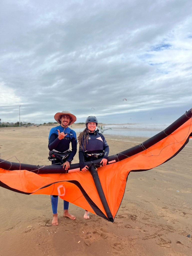 Escuela de kitesurf en Essaouira — pareja en la arena con ala naranja, neopreno y ambiente de playa.