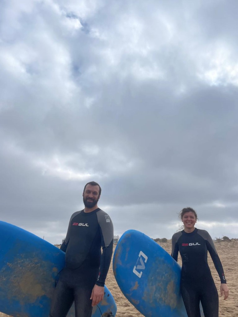 Pareja sonriente en neopreno en la playa con tablas azul soft — clases surf Essaouira.