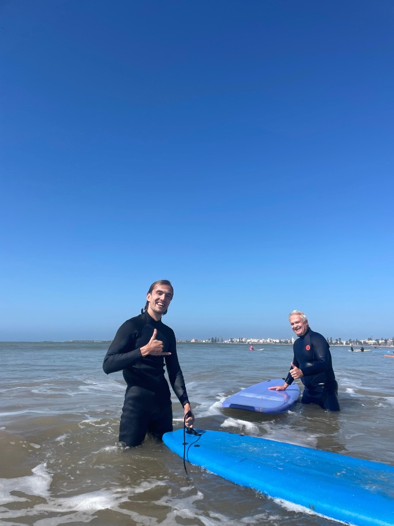 Dos surfistas en el mar con shaka, tablas azul y violeta — ambiente clase surf Essaouira.