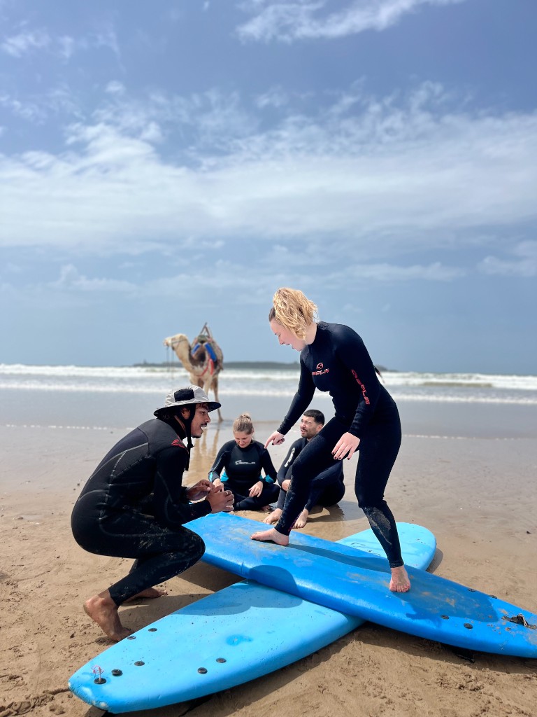 Clase de surf en la playa en Essaouira: monitor en neopreno corrige la postura sobre tabla azul, alumna practicando en la arena, mar y cielo al fondo