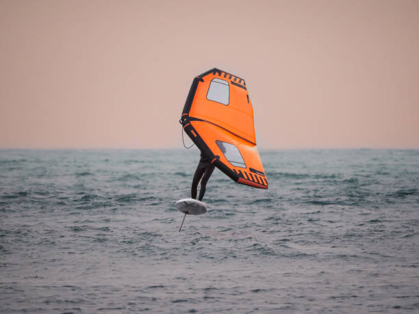 Wingfoilista en el aire sobre mar picada, neopreno negro, ala naranja con ventanas, tabla foil blanca y mástil negro — cielo melocotón uniforme, sesión en Essaouira.