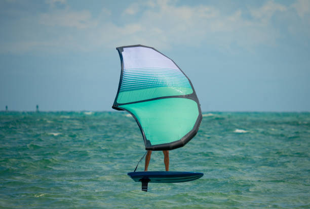 Wingfoil en mar turquesa — ala grande blanca y teal a rayas, tabla foil azul oscuro, cielo nublado.